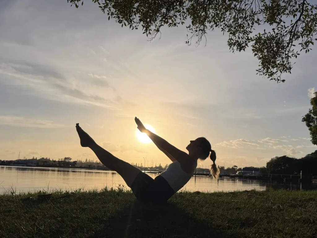 Woman performing Pilates teaser pose outdoors by the water at sunrise